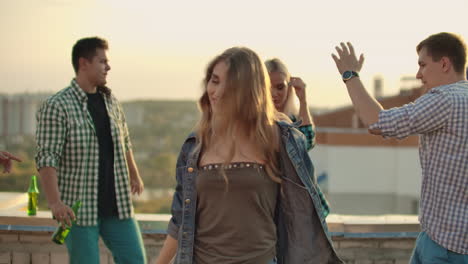 The-young-beautiful-American-girl-is-dancing-on-the-roof-with-her-four-friends-on-the-party.-She-smiles-and-enjoys-the-time-in-shorts-and-a-light-denim-jacket-in-summer-evening.-Her-blond-hair-is-flying-on-the-wind.